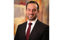 Joseph L. Albano, DO, MBA, smiles in black suit and red tie in front of bright background. 